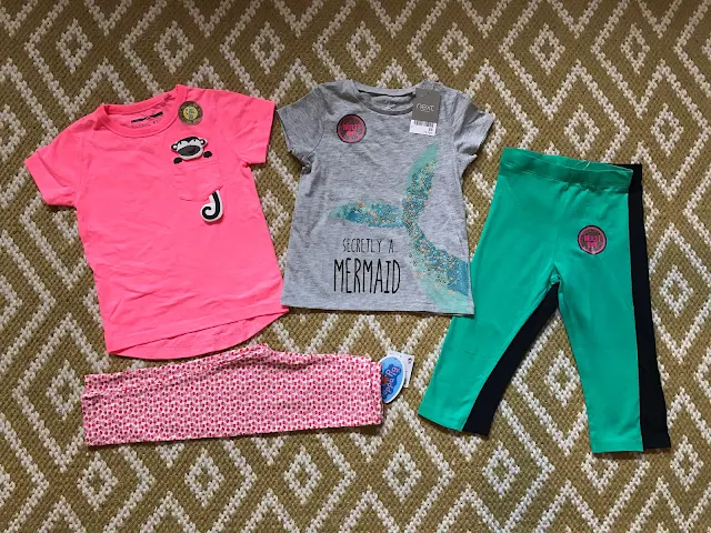 A selection of Next clothes on a rug: a pink "boys" t-shirt with a monkey on the breast pocket, a pair of leggings with a white background and lots of little peppa pigs, a grey t-shirt with a fishes tail and text saying "secretly a mermaid" and a two pack of leggings (green and navy) 