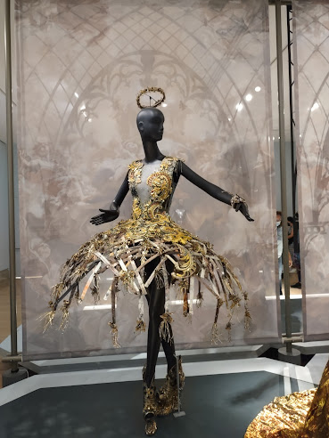 Photo: Guo Pei exhibit. Mannequin wearing brilliant, complex, gold platform shoes, a gold dress that is more of the frame of a dress than a garment,  including a flared skirt more or less at hip height and a sparse bodice, and a gold fascinator.
