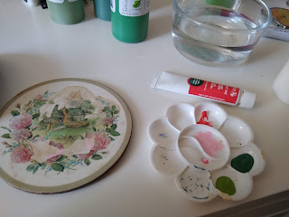 A damaged coaster that shows an image of an English cottage surrounded by roses sits on a table with a paint tray, paints and water.