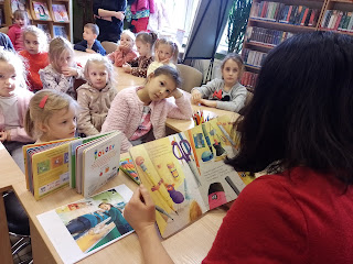 Pani bibliotekarka siedzi przed grupą dzieci i czyta im książkę.