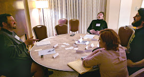 four people in a semi-circle around a table filled with index cards, paper, and writing instruments.
