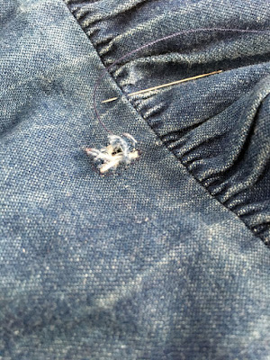 A close-up of a frayed hole in a piece of denim, just above a gathered seam showing wear on the tops of the gathered section, with a silver needle threaded with blue-purple sewing thread stabbed through the seam.