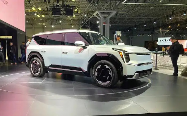 The Kia EV9 boxy, light-colored SUV is shown with lights in the background.