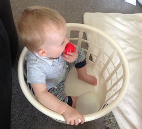Baby sitting in washing basket licking a red toy ball