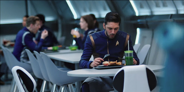  vemos a Sarek e uniforme comiendo solo en una mesa,una hamburguesa con cuchillo y tenedor,  con un batido muy verde con pajita  