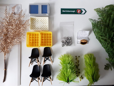 Flat lay of one-twelfth scale modern miniatures including Eames chairs, crates, a trolley and some fake greenery.