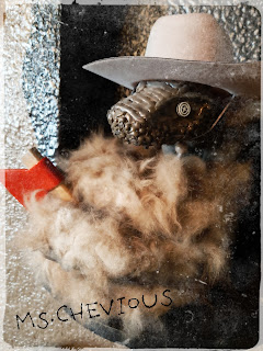 A photograph of "Snakesquatch," a mixed media sculpture of a snake with a fur covered body, wearing a hat and holding a tiny toy hatchet.