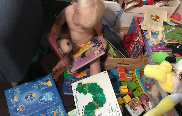 My toddler sitting on the floor reading a book surrounded by lots of books and toys