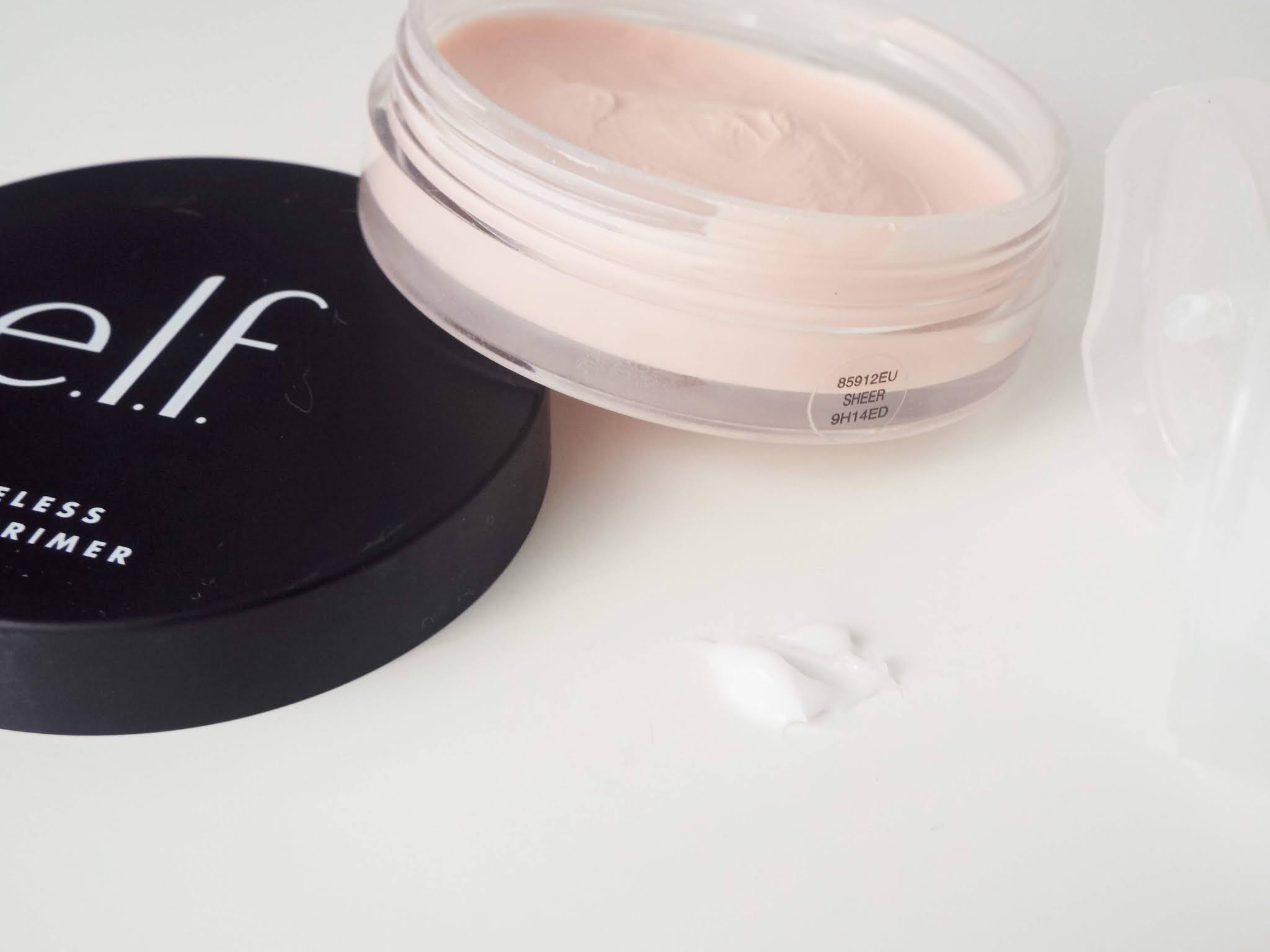 Close up shot of the Elf Poreless Putty Primer, open with lid resting just aside, revealing the light pink putty in pot, with lines from scooping product out. A blob of the opaque Ordinary High-Adherence Silicone primer sits in front, with a smooth but thick consistency.
