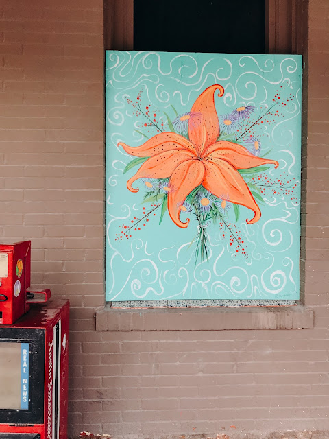 Orange flower painting sitting in window next to newspaper stand.