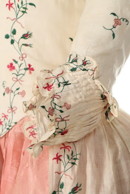 A detail of the sleeve cuff, with the bodice and petticoat visible in the background, showing the tiny green vine with a variety of pink carnations, rosebuds, and five-petalled flowers tied to the main vine at intervals.