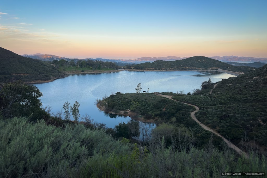 A still Lake Poway surrounded by green hilsides with distant mountains. The foreground is dark, but the distant mountains are shining from the early morning sun.