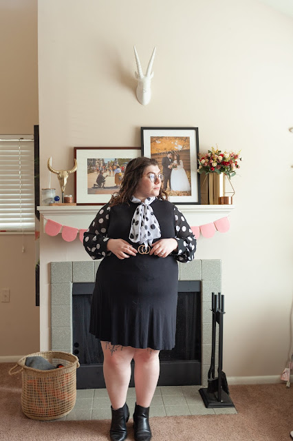 An outfit consisting of a white on black long sleeve blouse with a black on white neck tie tied in a bow under a sleeveless black knee length dress and black Chelsea boots