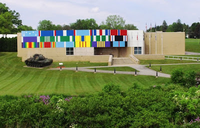 Front of PA Military Museum with array of simulated military ribbons that takes up the upper left two-thirds of the building. A row of service flags is to the right and a military tank is parked in front of the ribbons.