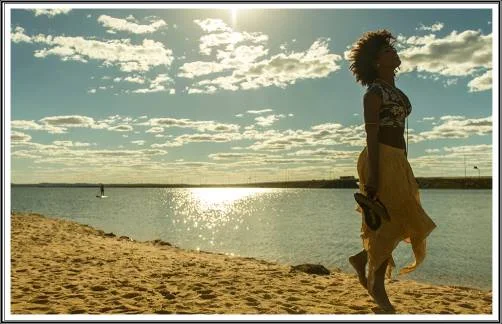 Paisagem de um grande LAGO, sol brilhante a iluminar, uma jovem pensativa, caminha descalça na areia.