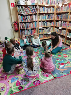 Biblioteka. Dzieci z bibliotekarką siedzą w okręgu. Za ich plecami regały z książkami.