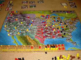 The game of Power Grid in the course of a four player game. The board represents a map of the continental United States with various cities connected by network lines. Wooden house tokens in the various players' colours have been placed on many of the cities. Along the bottom of the board are black, brown, orange, and yellow tokens representing the various resources. Cards and more tokens lie around the board on the table.