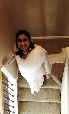 A young Indian woman in a pale pink sari and blouse with long, sheer sleeves, standing on beige carpeted stairs looking up at the photographer. She's grinning broadly, with her right hand up to brush her shoulder-length hair forward, and wearing a pearl and diamond necklace and earrings. The pallu trails behind her on the landing.