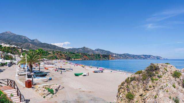 Playa con barcas y gente, un paseo marítimo con palmeras a su espalda, montañas al fondo y el mar a su frente.