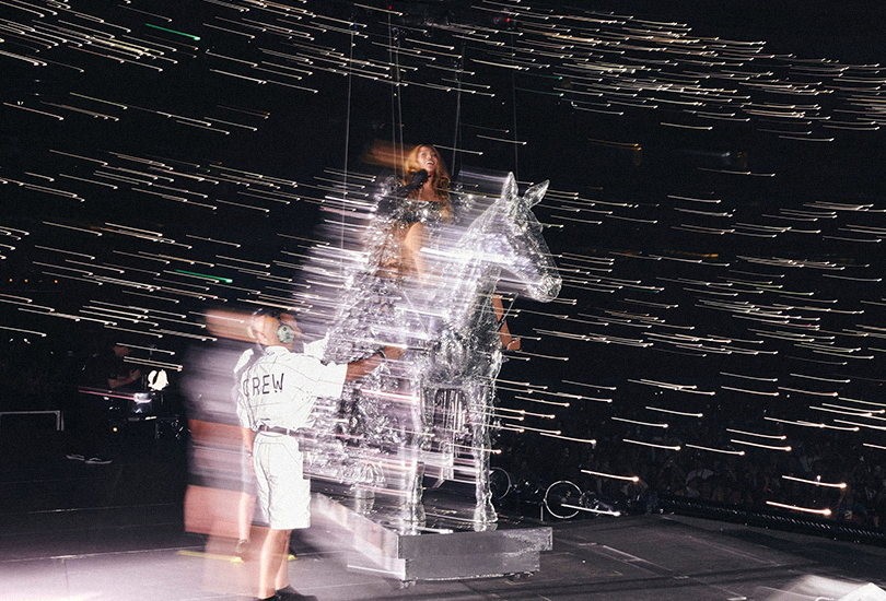 A shot of Beyoncé performing on the Renaissance World Tour. Beyoncé is sat atop the disco horse, as the stage crew assist.