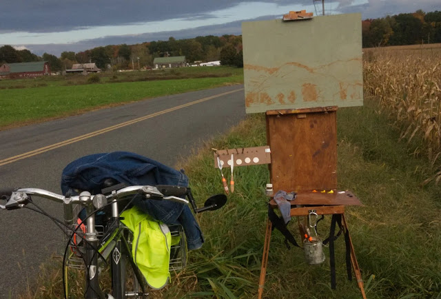 Bicycle and French easel with sketched in painting by side of country road with farm houses in background.