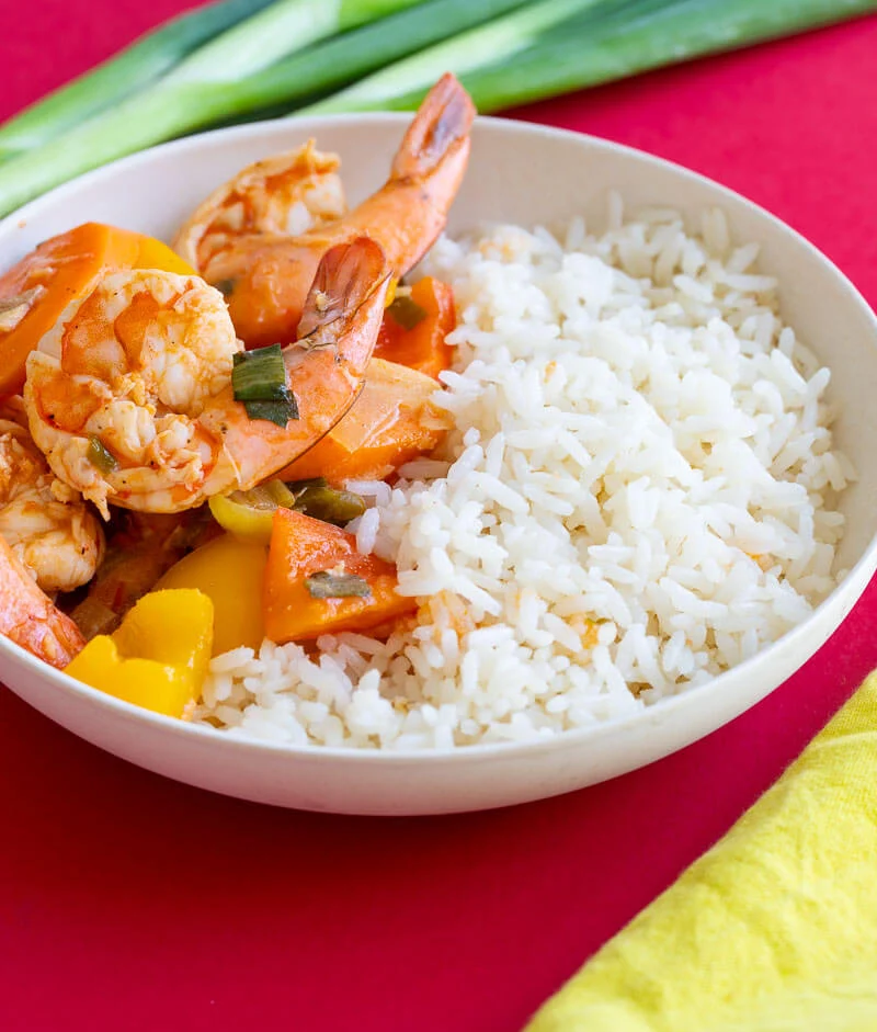 A bowl of pepper shrimp and coconut rice on top a red counter with a yellow napkin.