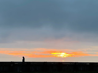 There's an orange setting sun amongst a blue sky. The beach is empty except for one person, silhouetted in black as she walks along.