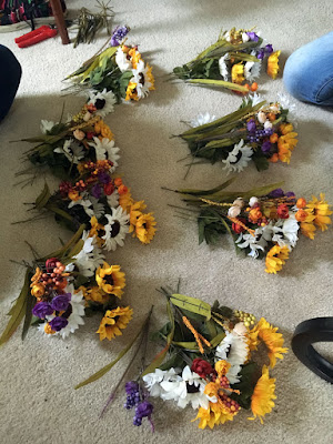 Eight small bundles of fake flowers, each composed of a mix of white and yellow sunflowers, small orange and purple roses, and red-to-yellow millet heads. Cut-off stems make a small, sad pile in the background, and two people's knees poke into the frame at opposite sides.