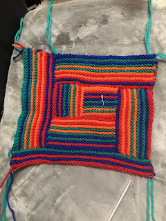 The piece has a decidedly rectangular shape currently, with the center square now having additional strips to the left side and bottom. The bottom strip, aka number 6, has 5 of its 12 garter stitch ridges complete.