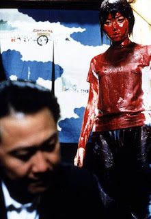 Standing girl covered in blood looking at seated man who ignores her