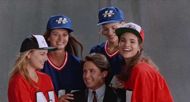 Gordon Bombay rodeado de mulheres lindas usando uniformes de hóquei. Ele tenta não parecer extremamente desconfortável com um sorriso amarelo.