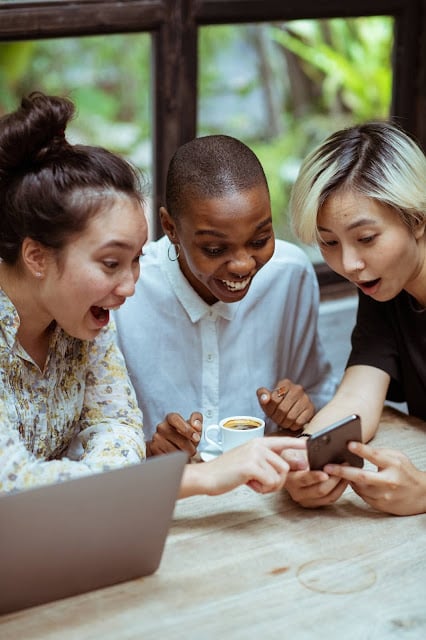 Happy Young Girls Looking Happily on Mobile Having Laptop and Coffee on Wooden Table