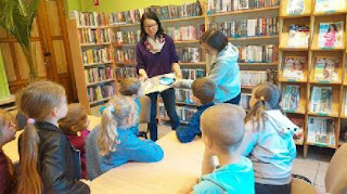 tło: regały z książkami. Panie bibliotekarki stoją przed grupka dzieci. Jedna z nich trzyma w ręku książkę a druga smartfona.