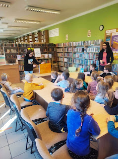 Pani bibliotekarka pokazuje dzieciom teatrzyk kamisibai. Dzieci siedzą naprzeciwko pani przy stolikach Tło: regały z książkami.