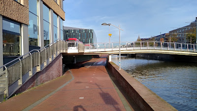 A cycle track dips under a bridge. It is next to a canal to the right and a building to the left. There is a little slip road off the cycle track into an entrance.