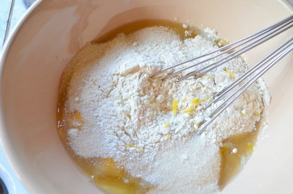 A cream colored mixing bowl with Eggs, Egg Yolks, Granulated Sugar, All-Purpose Flour, and Lemon Zest in it with a flat whisk resting on top.