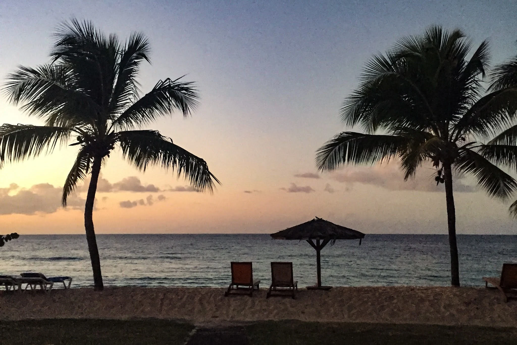 A sunset on a beach in Grenada showing palm trees an umbrella and seats