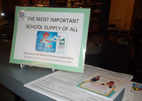 Sign on counter-top reads, 'The Most Important School Supply of All. Sign up for a free library card at your public library.' Below, it displays information for Ashland Branch Library, 410 Siskiyou Blvd., Children's Department: 541-774-6995