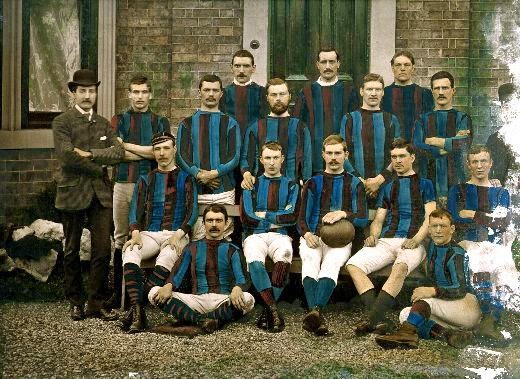 Photo of Castleford Football Club in 1884. The team are all young, white men, many with moustaches. Their manager stands to the left, in a suit and bowler hat. The club's colours of blue and magenta vertical stripes have been hand painted on.