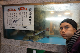 Pili haciendo de pez fugu con la boca hinchada. Detrás tiene una pecera en un restaurante en el que ofrecen pez globo. 