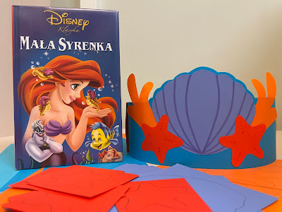 Na pierwszym planie papierowe komponenty potrzebne do wykonania pracy plastycznej. Dalej, po lewej stronie, stoi pionowo książka Walta Disney'a "Mała Syrenka", obok korona Arielki - opaska na głowę z muszlą, koralowcami i rozgwiazdami.