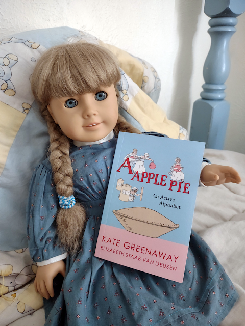 An American Girl Kirsten doll with blonde braids and a blue flowered dress holds a small book. The book is A Apple Pie: An Active Alphabet, by Kate Greenaway and Elizabeth Staab Van Deusen. The cover is blue and pink, with an illustration of four young children with baking supplies and a large pie.