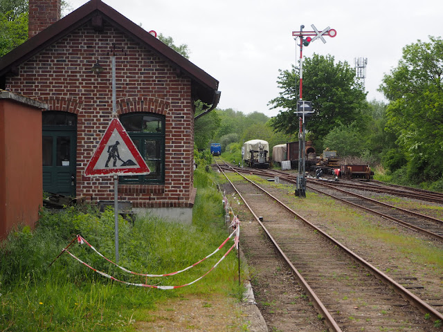 Kleines rotes Gebäude mit grüner Tür und Fenster. Davor ein Baustellenschild und hoher Rasen. Rechts daneben Gleise und ein Signalmast mit Flügelsignal. Im Hintergrund Waggons.