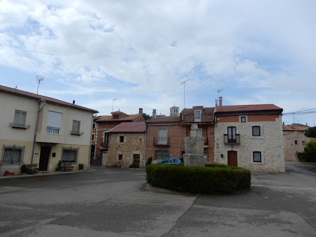 plaza con un monumento en medio hecho con dos piedras de lagar, rodeado por un jardincillo y rematado en una cruz. Al fondo se ve la torre de la iglesiat