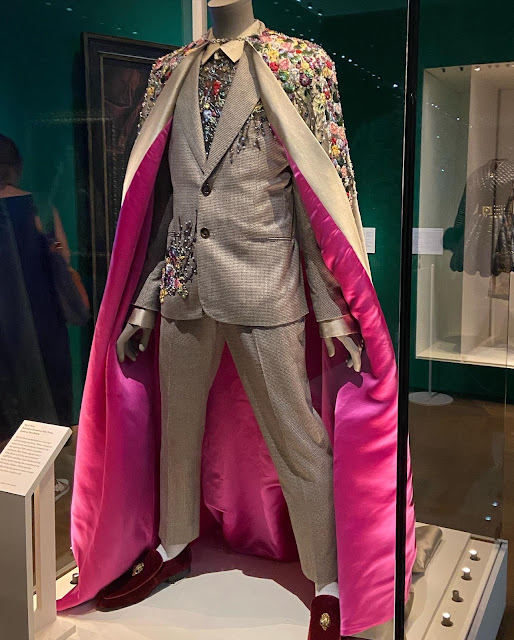 A grey suit and cape. The cape has a shocking pink lining, and there is floral embroidery on the shoulders of the cape, the front of the jacket, and thickly encrusting the shirt beneath.