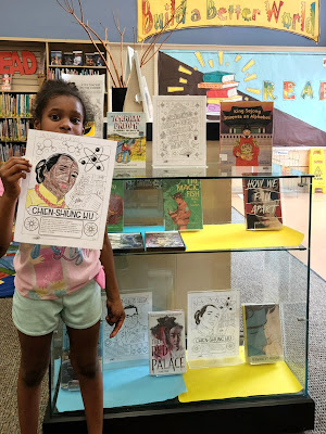 A young black girl holds up a coloring page she colored of Chien-Shiung Wu next to a AAPI Heritage Month display