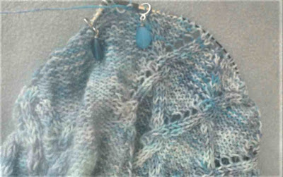 a cables and lace lace pattern with a background of reverse stocking stitch; worked in variegated blue and grey yarn.  A stitch marker with a blue sea glass charm is looped over the tip of the needles.