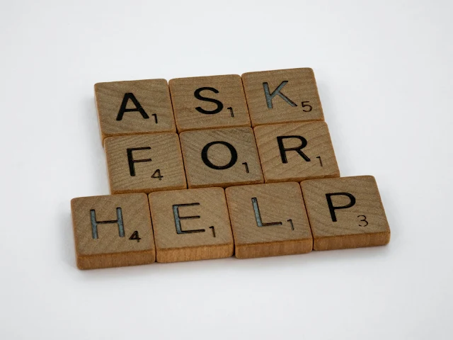 A set of wooden Scrabble tiles rest on a white surface. They are arranged to spell out the phrase "ask for help".