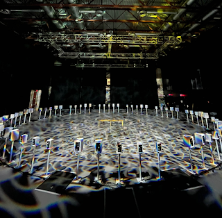 Photo of a large room with a mottled floor and ceiling and a gigantic circle of loudspeakers, each on an individual stand.