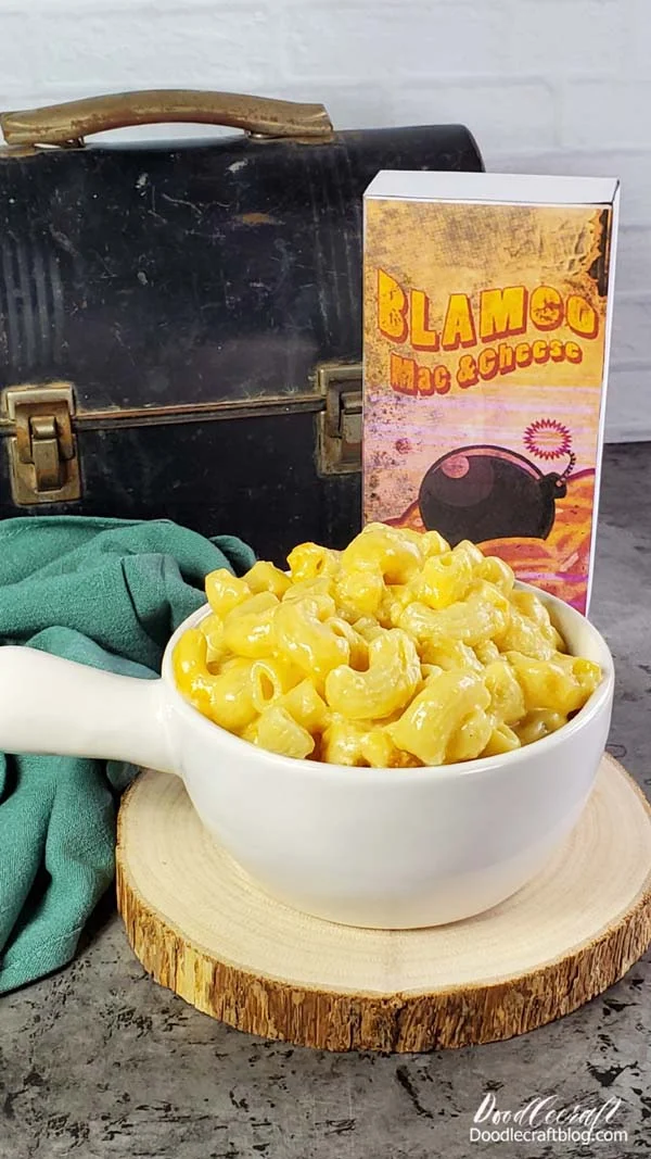 Blamco Mac and Cheese box sitting next to a dusty mole miner pail and a bowl of fresh homemade Macaroni and Cheese. Perfect for the Fallout Wastelander.